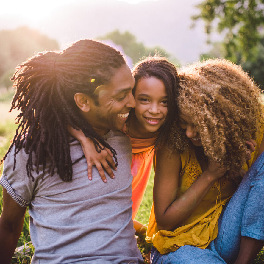 Soft glowing sunny moment between a young african-american father and his cute little girl and loving wife all giving eachother a big hug and smiling happily